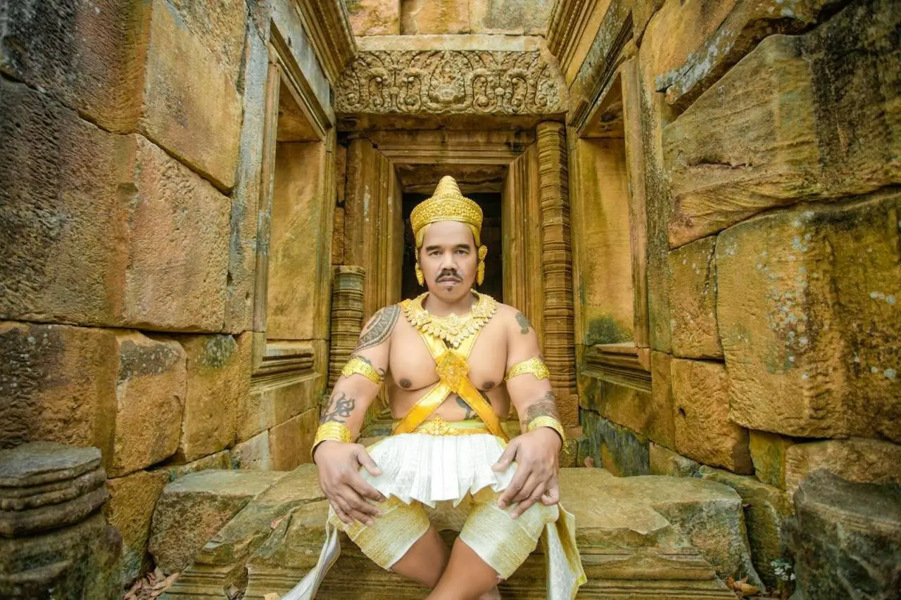 Man in golden traditional attire sitting at an ancient temple with intricate stone walls.