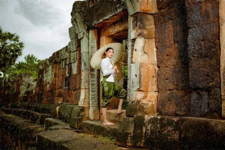 A woman elegantly poses in traditional attire at an ancient Cambodian temple, holding a parasol.