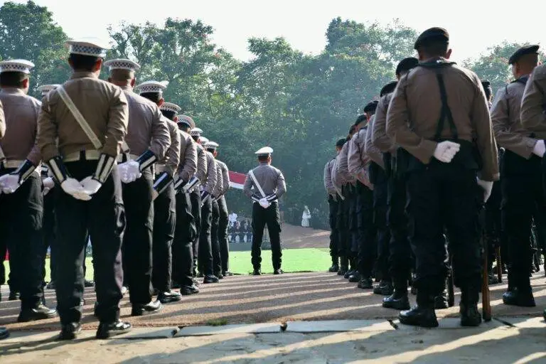 A group of police officers stands in formation outdoors, observed from behind, during a formal event.